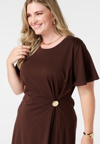 Robe t-shirt marron à manches courtes, avec un détail de taille torsadé et une accentuation en bouton doré. Tissu en coton lisse.