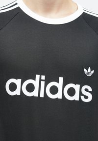 Czarna koszulka treningowa z krótkim rękawem, z białym kołnierzykiem i dużym białym napisem "adidas"; posiada kultowe białe logo trefoil po lewej stronie.