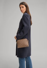Donna in cappotto blu navy e jeans blu porta una borsa a tracolla marrone con motivo e strap nero, in piedi contro uno sfondo grigio semplice.