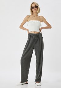 Wit smocked strapless top met een gerimpelde rand en donkergrijze high-waisted wijde broek. Model draagt witte instappers en een zonnebril.