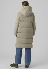Abrigo acolchado de un beige claro con capucha, presenta un diseño de acolchado y mangas largas. Se lleva sobre unos pantalones vaqueros de pierna ancha en azul y calzado oscuro.