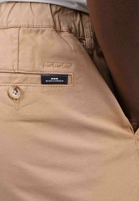 Pantalon beige au toucher lisse, avec une poche arrière à fermeture boutonnée et une étiquette de logo "MISSEAUGREEN" au-dessus de la poche.