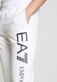 Λευκά φούτερ παντελόνια με χαλαρή εφαρμογή, διαθέτουν πλαϊνή τσέπη και μαύρα γραφικά του "EA" και "EMPORIO" στο αριστερό πόδι.
