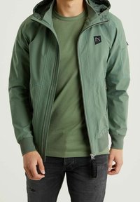 Green lightweight jacket with a zip front, hood, and mesh accents. Long sleeves and a subtle logo detail on the chest; paired with a green t-shirt.