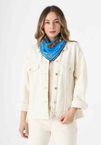 Lichtbruine denimjack met zilveren knopen, gedragen over een beige top. Een blauwe zijden sjaal met bloemachtige en Beierse ontwerpen is om de nek geknoopt.