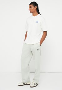 Vīrietis ar bizēm, ģērbies baltā Adidas T-kreklā, gaiši pelēkos Adidas sporta biksēs un baltās kedās ar melnām svītrām, stāv uz baltā fona.