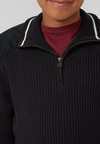 Schwarzer Reißverschluss-Pullover mit strukturiertem Strickmuster, weißen gestreiften Kragenakzenten und einem weinroten Hemd darunter. Reißverschluss am Hals.