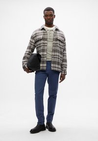 Grau kariertes Flanellhemd, das über einem hellgrauen Oberteil getragen wird, kombiniert mit blauen Hosen und schwarzen Lederschuhen. Hält in einer Hand eine schwarze Tasche.