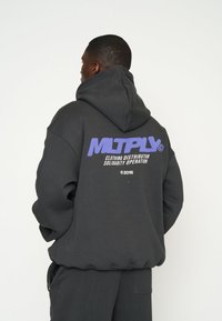 Felpa oversize grigio scuro con un grande logo viola sulla schiena, che presenta il testo: "MLTPLX CLOTHING DISTRIBUTOR SOLIDARITY OPERATION 2015."