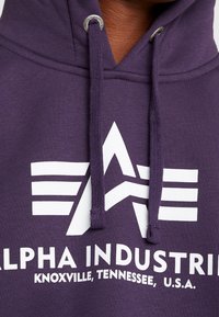 Gros plan d’un sweat à capuche violet avec le logo blanc Alpha Industries et le texte « Knoxville, Tennessee, U.S.A. » sur la poitrine, ainsi que des cordons avec embouts métalliques.