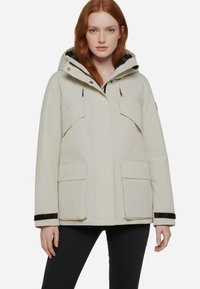 Giacca impermeabile beige con cappuccio, dotata di due tasche sul petto e due tasche laterali, dettagli neri sui polsini e una vestibilità morbida.