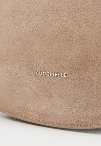 Smeđi sueded materijal s mekanom teksturom; sadrži srebrni logo "COCCINELLE" istaknut na vidnom mjestu. Oblik je zaobljen s urednim šavovima.
