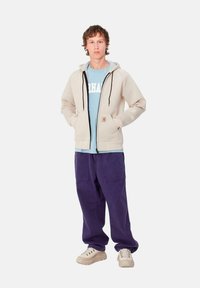 Sudadera beige con cremallera y cordones negros, superpuesta a una camiseta de color azul claro. Combinada con pantalones cargo morados y zapatillas beige.