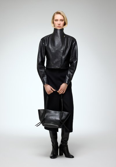 Černá kožená zkrácená bunda s vysokým límcem, doplněna černými šaty. Modelka drží černou koženou kabelku a má na sobě černé kozačky po kolena.