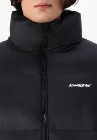 Svart puffer-jakke med høy krage, frontglidelås og en liten hvit logo. Glatt, lett stoff med quiltet tekstur.