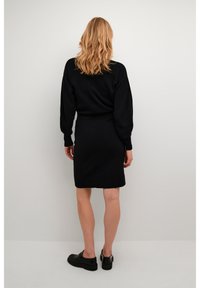 Femme blonde de dos, portant une robe noire à manches longues et des mocassins noirs, devant un fond clair.