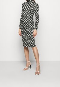 Czarna i biała sukienka w kratę z długimi rękawami i wysokim kołnierzem. Dopasowana sylwetka, dół do kolan. Modelka w białych sandałach na obcasie.