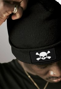 Black knit beanie featuring a flat patch with a white skull and crossbones design. The material shows a ribbed texture and snug fit.