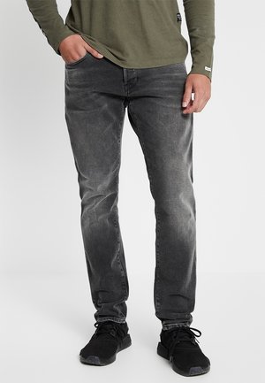 Jeans de hombre en denim negro con un acabado desgastado, diseño de pierna recta, cinco bolsillos y detalles de costura sutiles. Combinados con zapatillas negras.