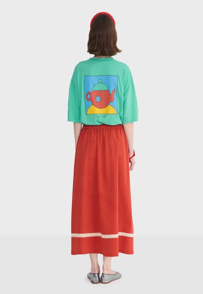 Femme de dos portant un T-shirt oversize turquoise avec un motif de théière, une longue jupe rouge avec bordure en dentelle, un bandeau rouge et des ballerines argentées.