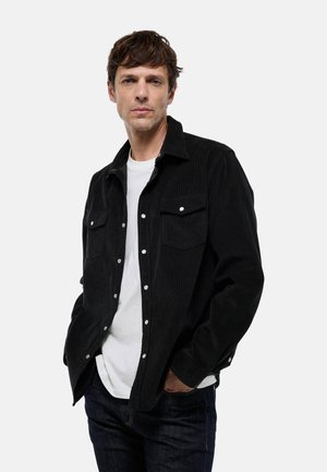 Chemise en velours côtelé noir avec des boutons-pression, deux poches poitrine, des manches longues et une coupe droite, portée sur un t-shirt blanc.