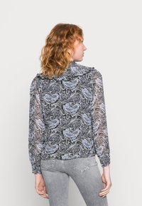 Blusa de manga longa em preto com um padrão paisley azul. Tecido transparente com um decote franzido e um ajuste descontraído. Usada com jeans cinzentos claros.