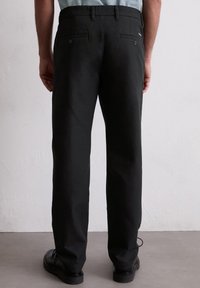 Schwarze Hose mit geraden Beinen, aus Baumwollmaterial, mit Knöpfen an den Gesäßtaschen und einer glatten Textur. Tragen Sie schwarze Schuhe und werden von hinten betrachtet.