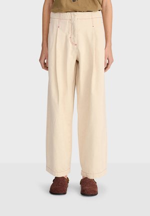 Pantalon beige à jambes larges avec coutures rouges, porté avec des chaussures marron texturées à enfiler et un haut marron, présenté de la taille aux pieds sur un fond clair.