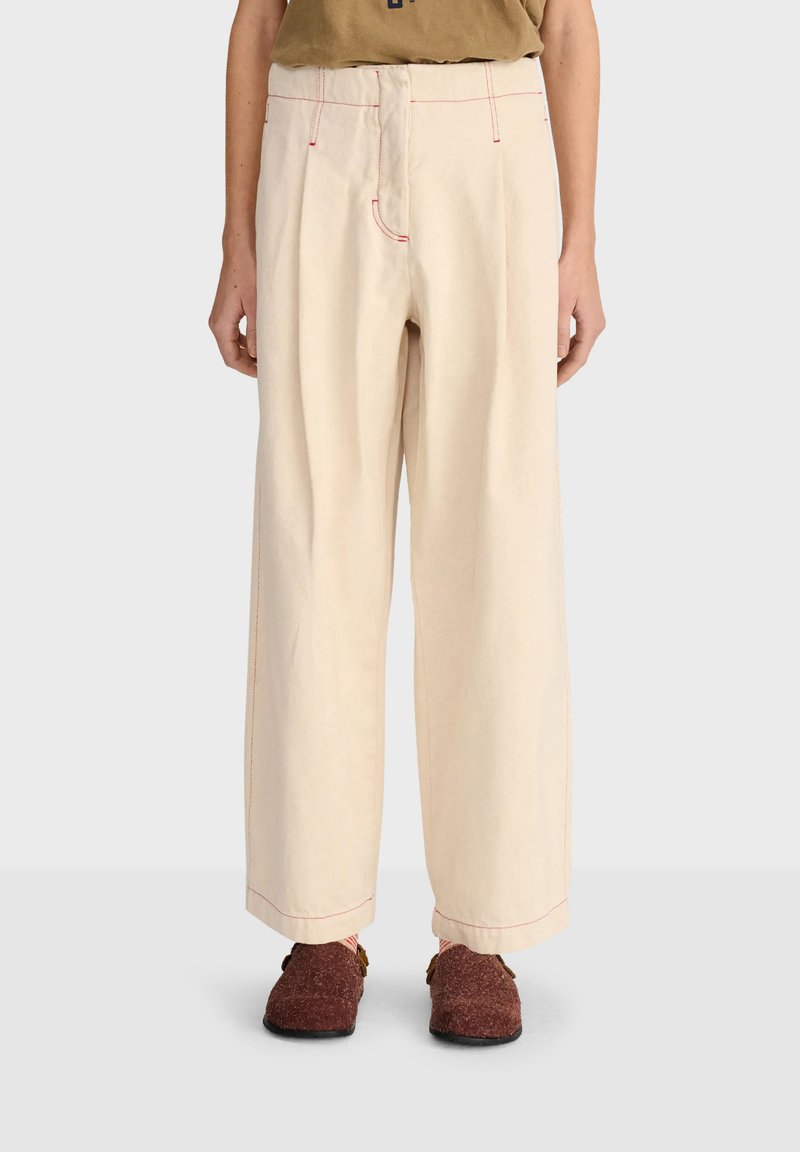 Pantalon beige à jambes larges avec coutures rouges, porté avec des chaussures marron texturées à enfiler et un haut marron, présenté de la taille aux pieds sur un fond clair.