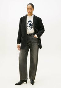 Zwarte textuur blazer over een witte sweatshirt met een blauw logo, gecombineerd met hooggetailleerde, wijde grijze jeans en puntige zwarte hakken.