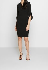 Svart klänning med nyckelhålsringning, vevade trekvartsärmar, figurnära silhuett och slätt tyg. Bärs med svarta klackskor med remmar.