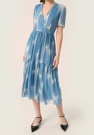 Woman wearing a blue and white striped midi dress with short sleeves, V-neck, and pleated skirt paired with black pointed-toe ankle-strap flats.