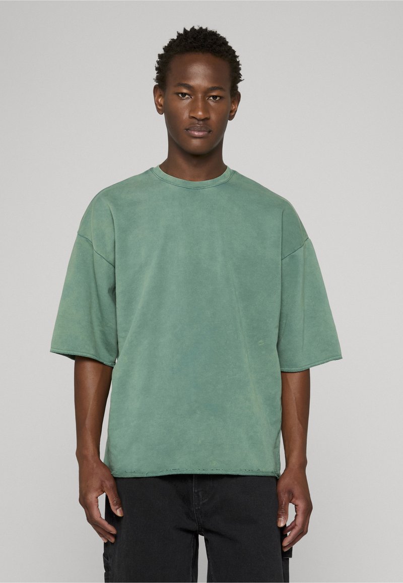 T-shirt oversize à manches courtes en coton vert délavé. Présente un col rond, des épaules tombantes et des coutures visibles le long de l'ourlet.