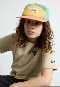 Adjustable cap in green, yellow, and orange with a front logo patch. Beige t-shirt with a logo and short sleeves. Curly hair visible.