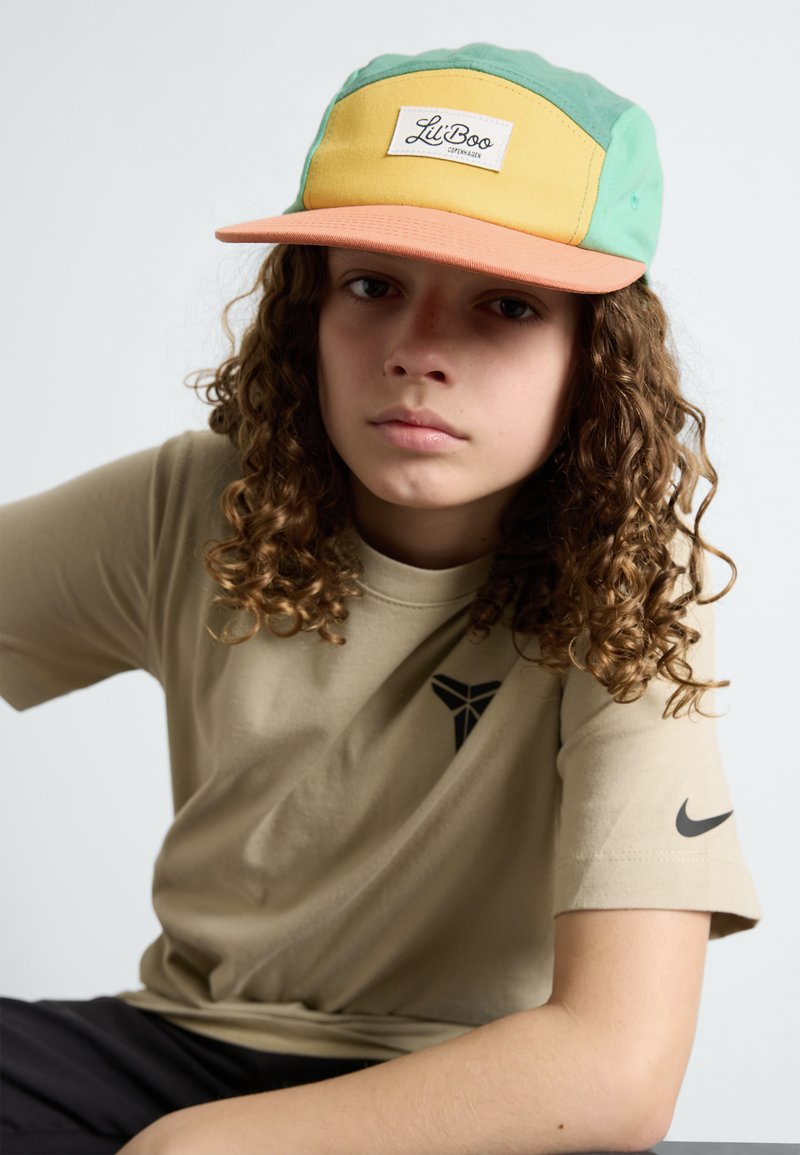 Adjustable cap in green, yellow, and orange with a front logo patch. Beige t-shirt with a logo and short sleeves. Curly hair visible.