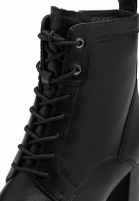 Bota de cuero negra hasta el tobillo con un cordón en la parte delantera, ojetes y un acabado texturizado. La suela es ligeramente elevada, con bordes estructurados.