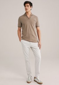 Beige polo shirt met korte mouwen, een V-hals en opgerolde mouwen, gecombineerd met witte broeken en groene sneakers met tan accenten.