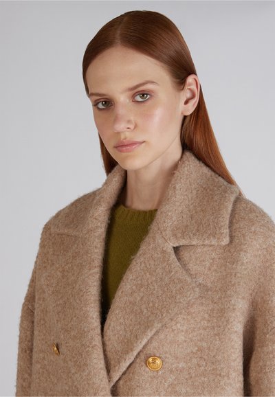 Podwójnie zapinany płaszcz z miękkiej beżowej wełny o strukturalnym wykończeniu, z dużymi klapami i guzikami w złotym odcieniu. Noszony na zielonym swetrze dzianinowym.