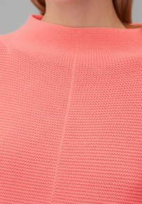 Korallefarbener Strickoberteil mit hohem Ausschnitt, aus strukturierter Stoffqualität und sichtbaren Nahtdetails, vor einem neutralen Hintergrund präsentiert.