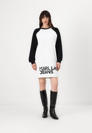 Rochie albă cu mâneci lungi negre și inscripția "KARL LAGERFELD JEANS" tipărită cu litere mari negre la tiv; modelul poartă cizme strălucitoare negre până la genunchi.