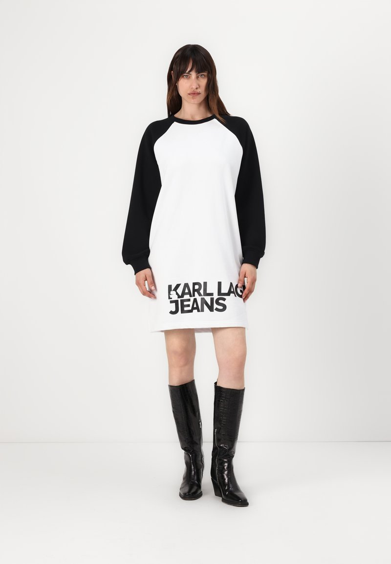 Vestido branco com mangas longas pretas e "KARL LAGERFELD JEANS" impresso em negrito preto na barra; modelo usa botas de joelho brilhantes em preto.