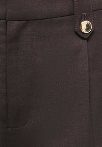 Gros plan d'un pantalon en tissu sombre montrant une poche avant et un bouton argenté brillant sur la ceinture.