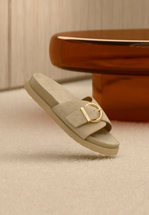 Sandale à glissière en daim beige avec une boucle circulaire dorée et une semelle texturée, posée sur une surface beige douce et une table orange.