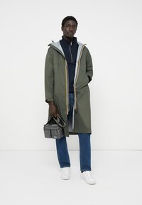 Giacca impermeabile verde con cappuccio, dotata di una fodera grigia, dettagli arancioni e una zip frontale. Indossata sopra una maglietta blu navy e jeans blu.