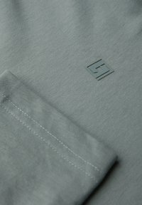 Harmaan puuvillakankaan sileä pinta, jossa on pieni kohokuvioitu logo kulmassa ja huomaamaton ompeluyksityiskohta helman varrella.