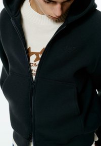 Felpa nera con zip in materiale felpato, dotata di una tasca frontale e un logo discreto. Indossata sopra un maglione di colore chiaro con un collo testurizzato.