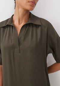 Dunkelgrünes Hemd mit Kragen, V-Ausschnitt und kurzen Ärmeln. Hergestellt aus einem glatten, leichten Stoff mit dezentem Glanz und minimalen Nähdetails.
