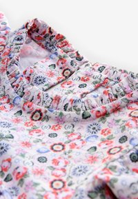 Nahaufnahme eines Stoffes mit floralem Muster aus kleinen roten, blauen und grünen Blumen, mit elastischen, gerafften Kanten und einem Knopfloch am Ausschnitt.