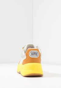 Sneaker features a mesh upper in white, orange leather accents, and a yellow outsole. Rear displays a "MAISON SCOTCH" logo on the heel.