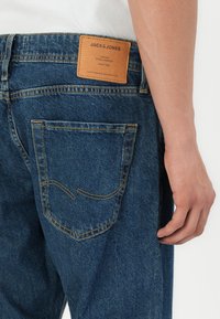 Vedere din spate a unei persoane care poartă blugi Jack & Jones de culoare albastră, cu o etichetă de piele vizibilă deasupra buzunarului din spate drept.